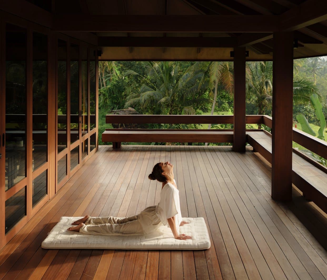 A woman arching up from a Heveya mat on the timber floor of an open pavilion, palms and jungle beyond the railing.